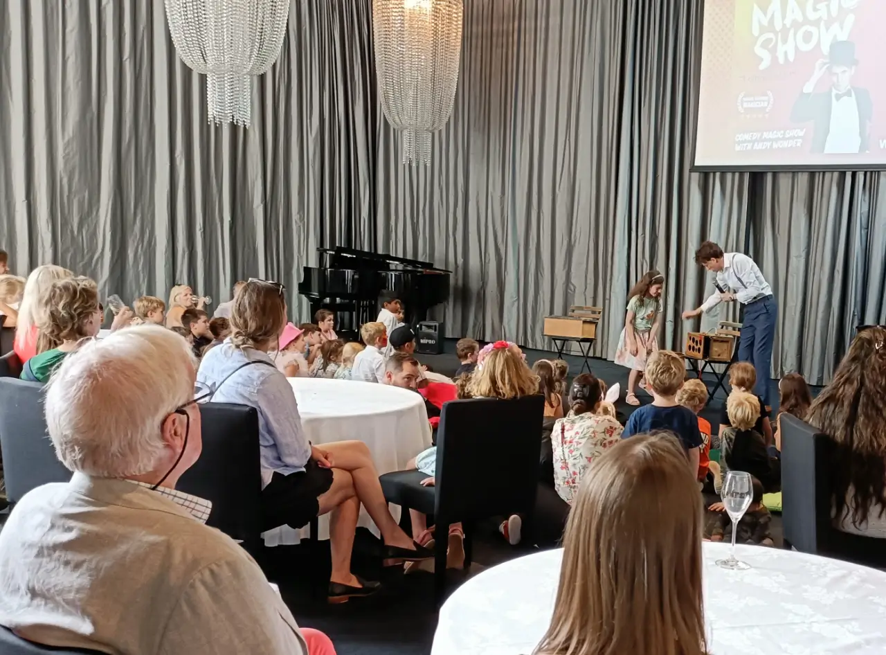 Elegant private theatre, grand piano, captivates New Zealand audience, children enchanted by magician below promotional screen.