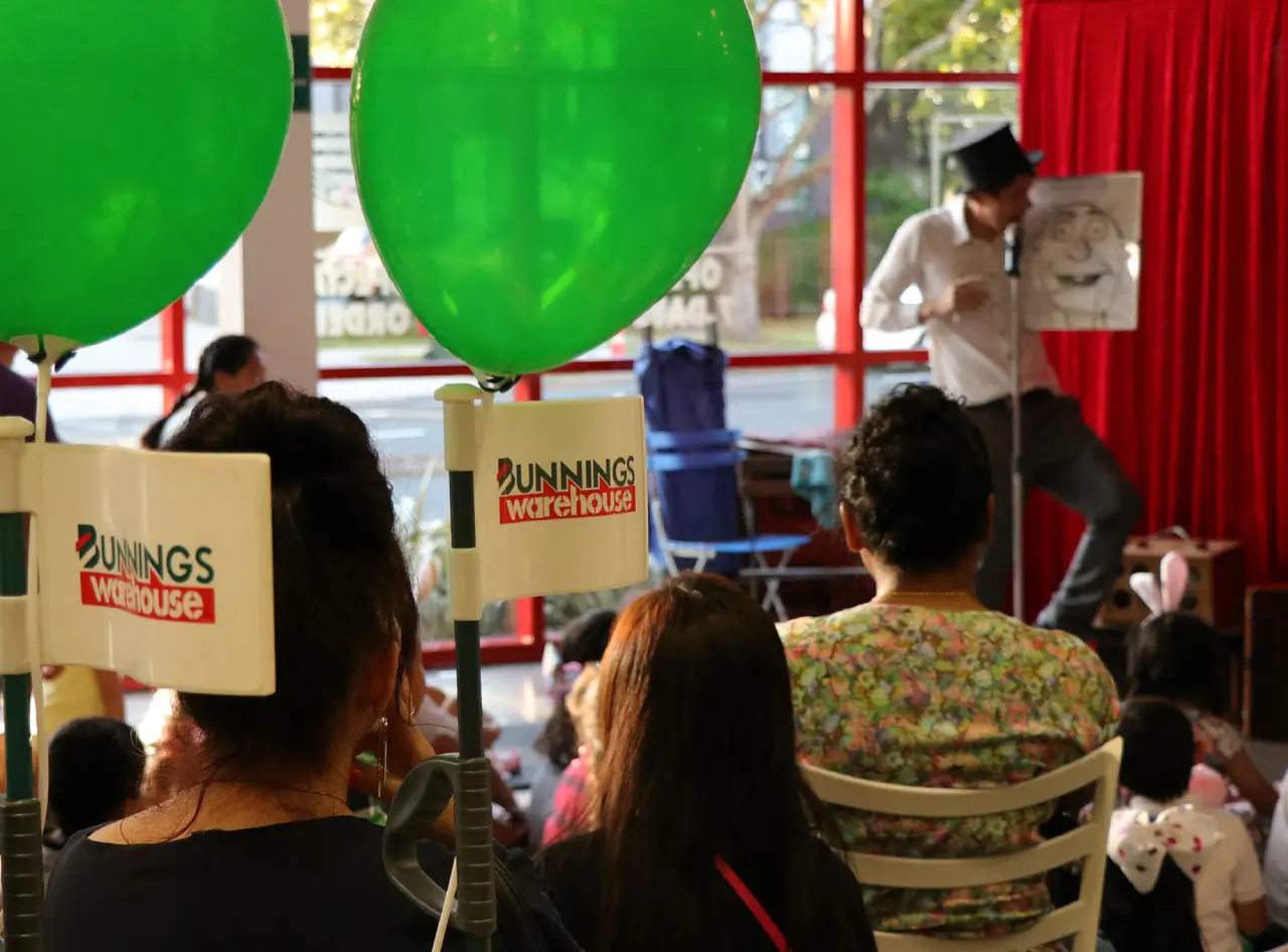 Crowd at Bunnings New Zealand relishes Andy Wonder's comedy magic amid green balloons, red curtain backdrop.